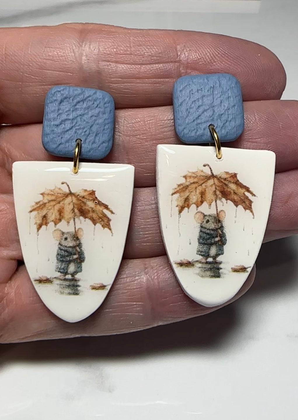 Polymer clay dangle earrings with blue textured studs and a whimsical mouse holding a leaf umbrella design on ivory background