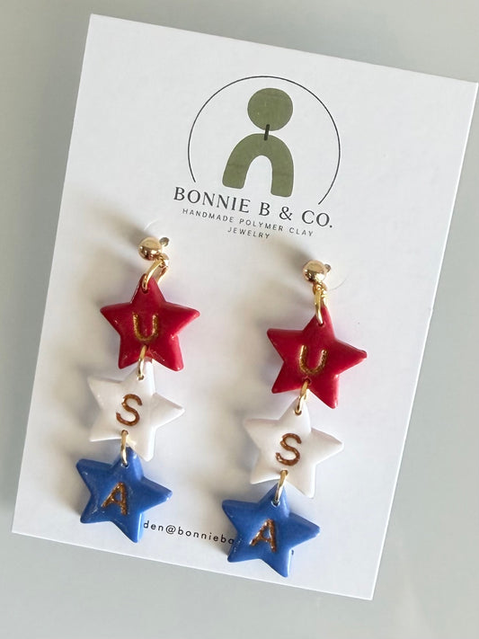 Handmade red white and blue patriotic USA star dangle earrings made from polymer clay with gold accents on a Bonnie B & Co display card.